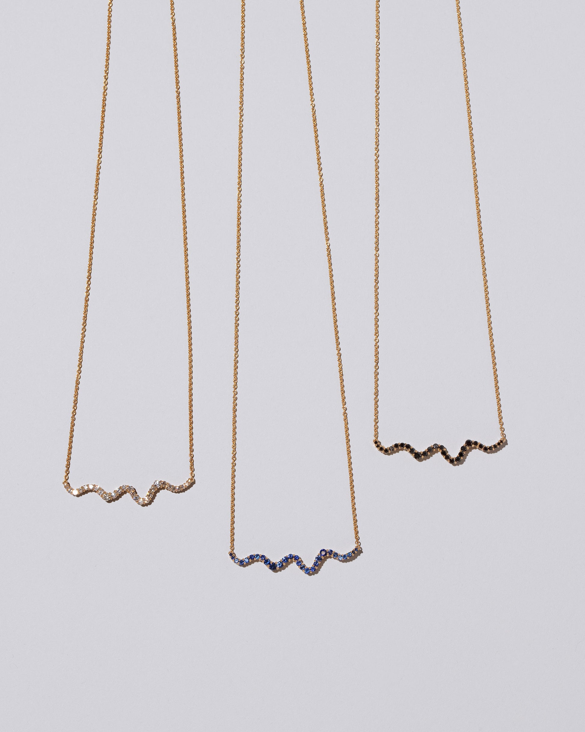 Closeup details of a group of Blue Sapphire, Black Diamond and White Diamond Ripple Wave Necklaces on light color background.
