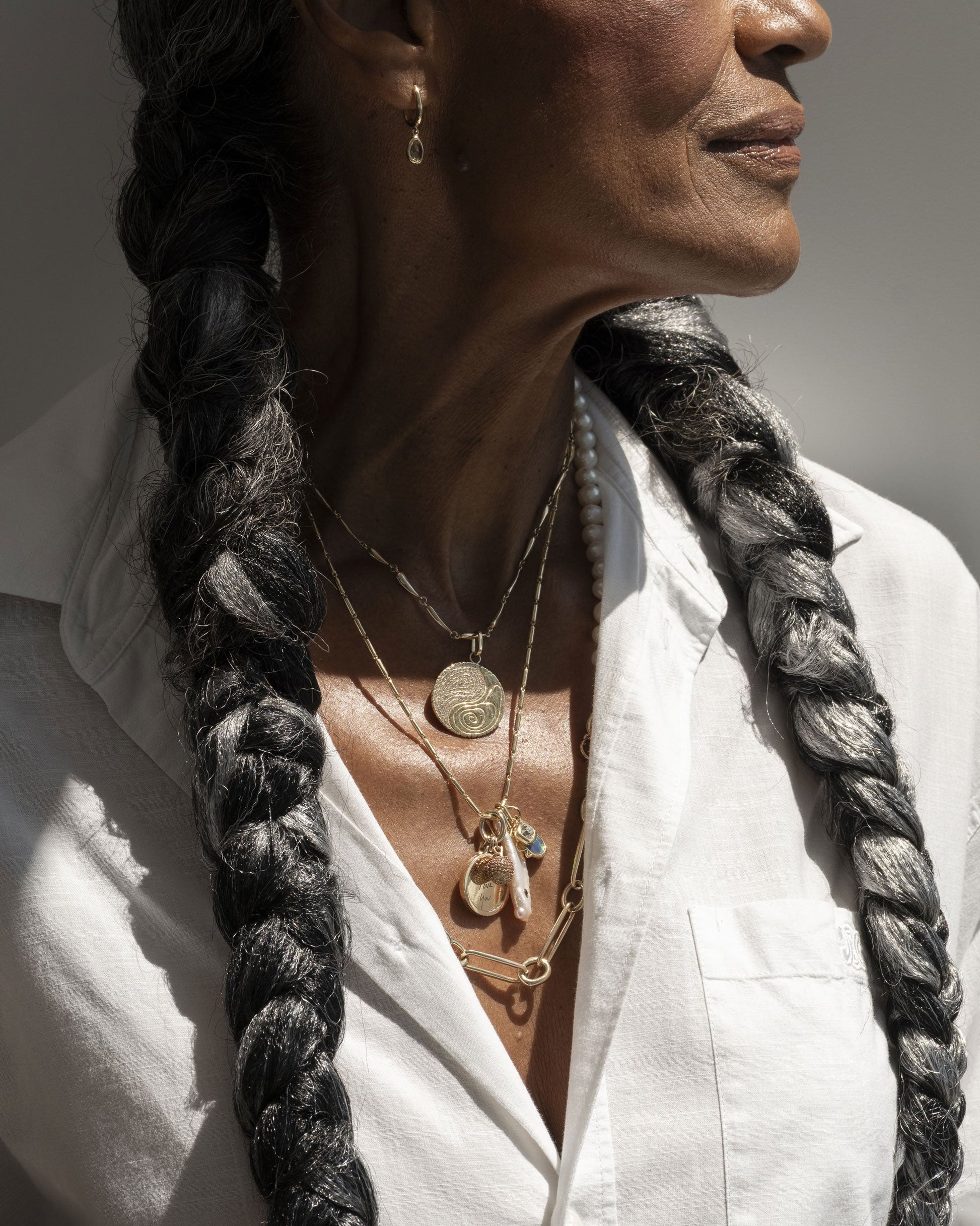 Styled image on model featuring the Yellow Sapphire Gusher Charm Huggie Hoop Earrings, Link Necklace, an Accumulation Necklace paired with the Grand Duality Medallion Pendant, and a Gold Oval Openable Link added to the Gold Butter Chain Necklace that holds a Birthstone Pearl Pendant, Peach Charm, Small Round Memento Pendant, Harmony Pendant and Diamond Poise Charm.
