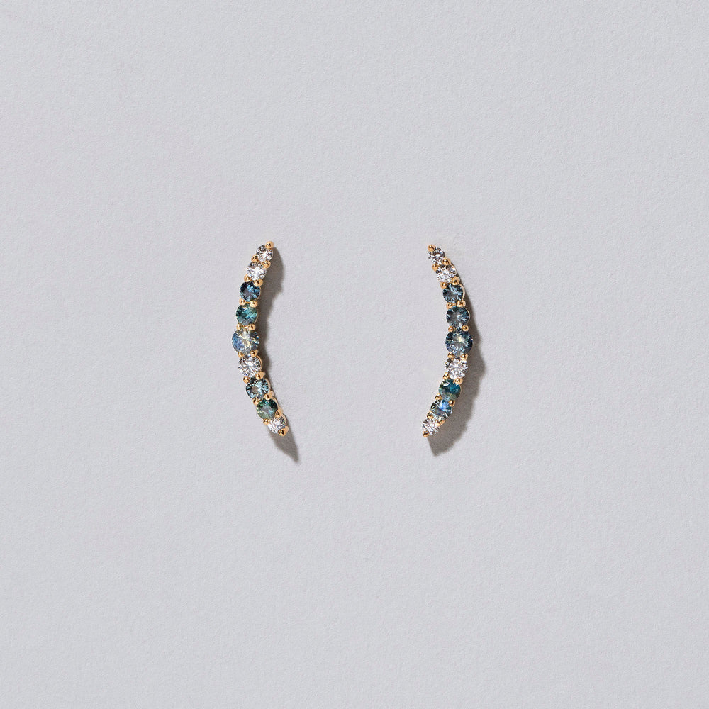 product_details::Closeup view of an edition of the Sapphire & White Diamond Crescent Ear Climber Stud Earrings on light colored background.