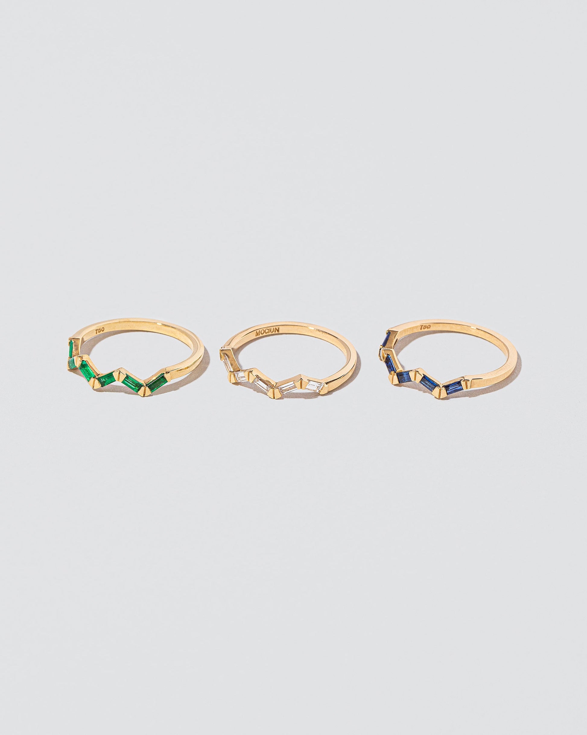 Closeup detail of Emerald, Sapphire and White Diamond Baguette Yellow Gold Zig Zag Bands on light color background.